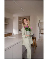 Een glimlachende vrouw met krullend haar, gekleed in een lichtgekleurd pak, staat in een moderne, minimalistische keuken met in haar handen een Kunstbloem Petunia UV 25 cm donkerroze met levendig roze bloemen.