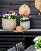 Twee grote keramische potten met een Kunstbloem-petunia UV 25 cm donkerroze, met levendige roze bloemen, staan op een tafel tegen een donkere houten muur. Op de tafel ligt een strohoed en boven de bloemen hangen gedroogde palmbladeren aan de muur.
