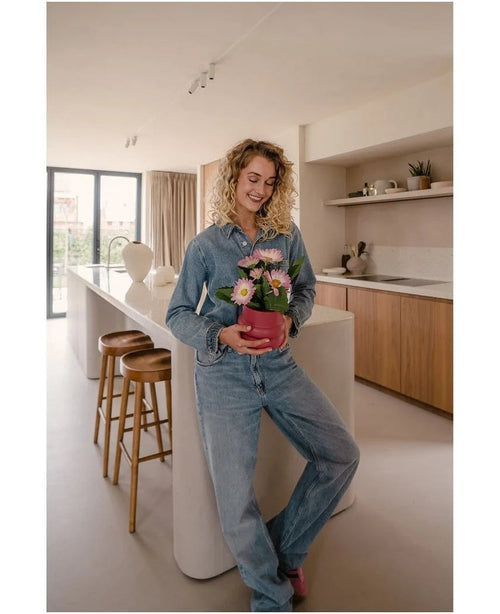 Une femme en jeans se tient dans une cuisine moderne, souriante et tenant dans ses mains la plante artificielle Gerbera UV rose 28 cm – un petit pot de fleur rose avec des fleurs roses et blanches. Au fond, on aperçoit deux tabourets en bois et une grande fenêtre.