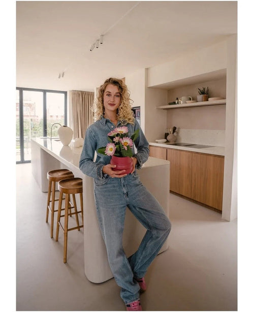 Une personne en tenue en jean se tient dans une cuisine moderne et sourit à la caméra tout en tenant la plante artificielle Gerbera UV 28 cm rose. Des tabourets en bois sont disposés autour de l’îlot de cuisine et des étagères décoratives sont visibles en arrière-plan.