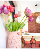 Een roze vaas met hartjes in reliëf houdt de Kunstbloem Franse Tulp 60 cm rood op een eettafel. Daarachter staan stoelen met rieten ruggen, een taart op tafel en een spiegel op een gedessineerde bruine muur onder een geweven hanglamp.