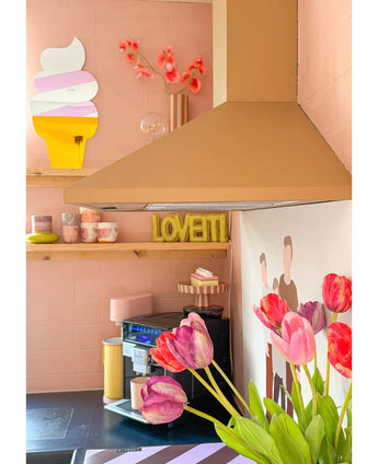 Een gezellige roze keuken heeft een Kunstbloem Franse Tulp 60 cm rood op de voorgrond, pastelkleurige kopjes op een plank, een LOVETTI bord, een muurschildering van twee mensen en een grillige ijshoornspiegel aan de muur.