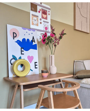 Een klein houten bureau met een stoel is versierd met Kunstbloem Catnip 75 cm roze, een mok, een gele ringsculptuur en kleurrijke kunstprints aan de muur erboven. Rechts is een bank gedeeltelijk zichtbaar.