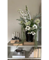 Sur une étagère contre un mur clair se trouve un vase noir avec la fleur artificielle Amaranthus 124 cm verte, à côté de cinq bougies blanches, des livres beiges et des objets décoratifs aux tons neutres.
