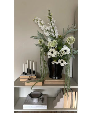 Sur une étagère contre un mur clair se trouve un vase noir avec la fleur artificielle Amaranthus 124 cm verte, à côté de cinq bougies blanches, des livres beiges et des objets décoratifs aux tons neutres.