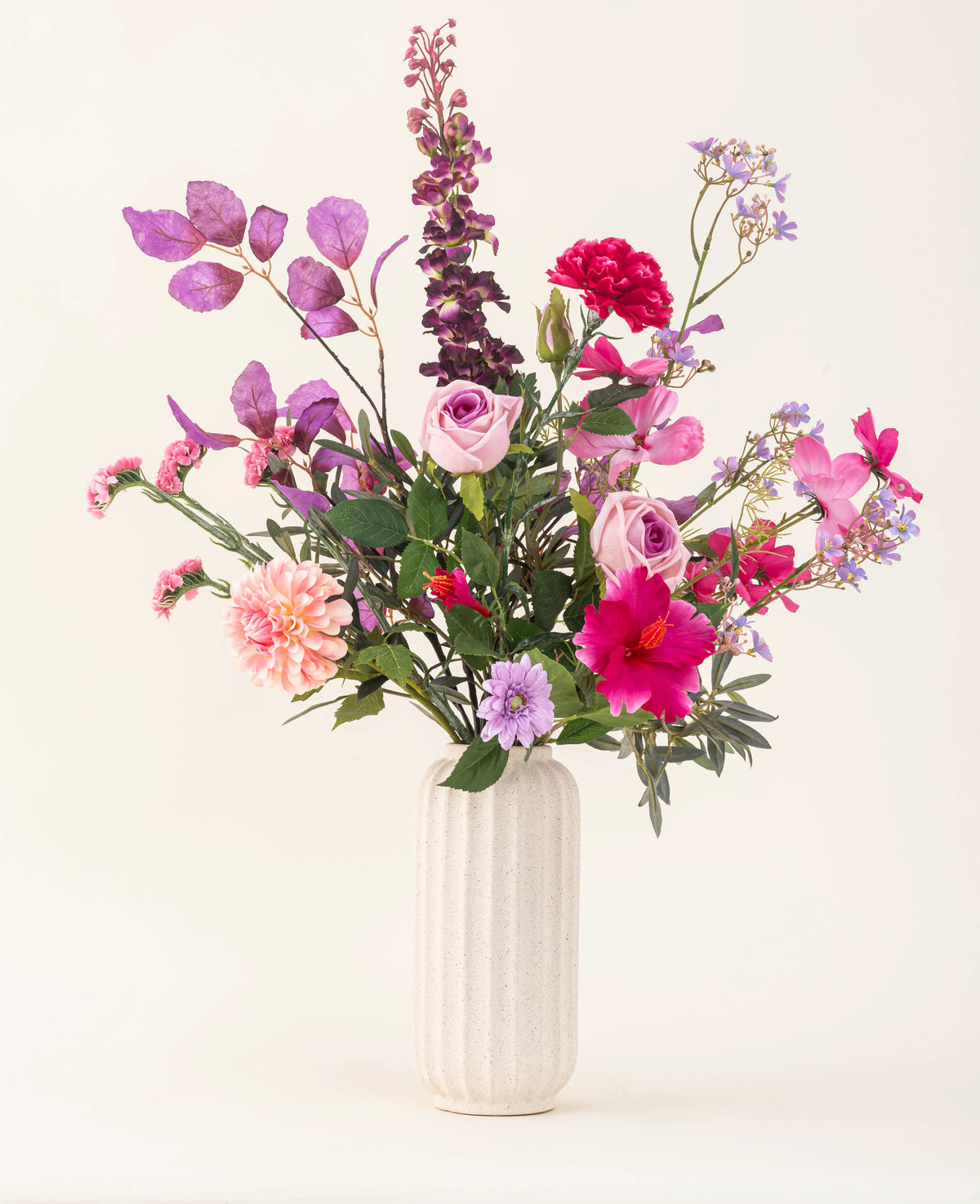 Un vase blanc texturé contient le bouquet de fleurs artificielles en soie Purple Dawn dans des tons rose, violet et lavande avec un feuillage vert, sur un fond clair et neutre.