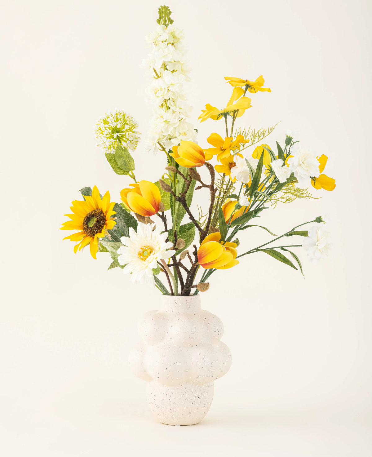 Le bouquet de fleurs artificielles en soie Sun Glow avec des tulipes jaunes, des tournesols, des marguerites et des feuilles vertes est placé dans un vase blanc structuré sur un fond uni blanc cassé.