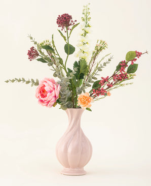 Het Zijden kunstbloemen boeket Pretty Petite, in een pastelroze vaas, bevat handgemaakte roze en oranje bloemen, witte bloemen, groene bladeren en rode bessen - perfect voor een duurzaam interieur tegen een effen lichte achtergrond.