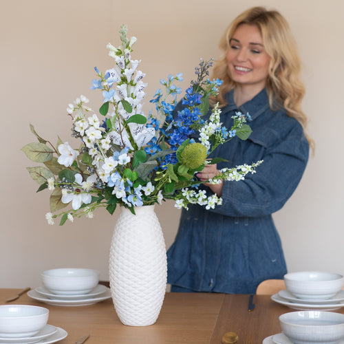 Une femme souriante vêtue d'une robe en jean bleue dispose le bouquet de fleurs artificielles en soie Cool Breeze dans un vase blanc structuré sur une table à manger garnie de vaisselle blanche et de couverts dorés.