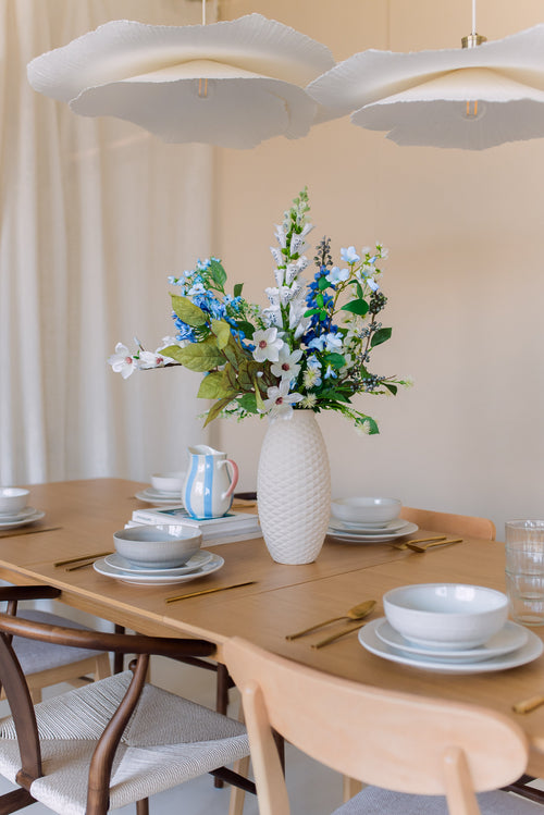 Une table à manger en bois recouverte d'assiettes, de bols, d'ustensiles de cuisine dorés et d'un vase blanc rempli du bouquet de fleurs artificielles en soie Cool Breeze. Un éclairage doux et des lampes suspendues blanches modernes au-dessus créent une atmosphère accueillante.