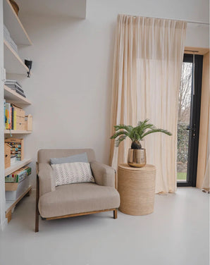 Un coin lecture cosy avec un fauteuil beige, une table d'appoint ronde en osier sur laquelle est posée la plante artificielle Baby Cycas Palm 60 cm, des bibliothèques et des voilages qui pendent au-dessus d'une porte vitrée qui s'ouvre sur le jardin.