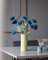Un coquelicot artificiel de 73 cm de couleur bleu foncé se dresse dans un vase jaune sur une table, accompagné d'une petite tasse à rayures colorées, dans une pièce moderne et doucement éclairée au décor minimaliste et aux tons neutres.