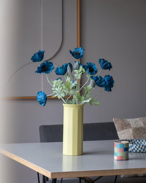Un coquelicot artificiel de 73 cm de couleur bleu foncé se dresse dans un vase jaune sur une table, accompagné d'une petite tasse à rayures colorées, dans une pièce moderne et doucement éclairée au décor minimaliste et aux tons neutres.