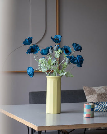 Un coquelicot artificiel de 73 cm de couleur bleu foncé se dresse dans un vase jaune sur une table, accompagné d'une petite tasse à rayures colorées, dans une pièce moderne et doucement éclairée au décor minimaliste et aux tons neutres.