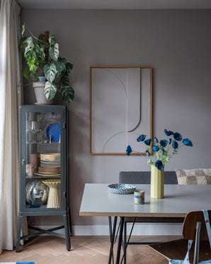 La salle à manger moderne comprend une vitrine en verre gris dans laquelle sont exposées de la vaisselle et des plantes, des décorations murales minimalistes et une table sur laquelle se trouve la fleur artificielle Coquelicot 73 cm bleu foncé dans un vase jaune. L'espace est complété par un mélange éclectique de chaises, le tout sur fond de murs gris et de parquet.