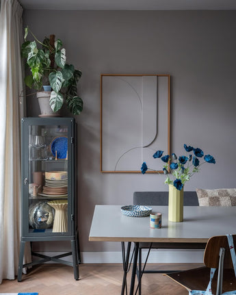 La salle à manger moderne comprend une vitrine en verre gris dans laquelle sont exposées de la vaisselle et des plantes, des décorations murales minimalistes et une table sur laquelle se trouve la fleur artificielle Coquelicot 73 cm bleu foncé dans un vase jaune. L'espace est complété par un mélange éclectique de chaises, le tout sur fond de murs gris et de parquet.