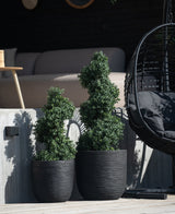 Deux plantes topiaires artificielles de style romain en spirale de buis de 90 cm avec des feuilles en spirale se dressent sur un patio en béton à côté d'une chaise suspendue noire et d'un canapé d'extérieur moderne en arrière-plan.