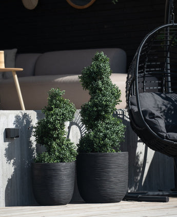 Deux plantes topiaires artificielles de style romain en spirale de buis de 90 cm avec des feuilles en spirale se dressent sur un patio en béton à côté d'une chaise suspendue noire et d'un canapé d'extérieur moderne en arrière-plan.
