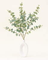 Een moderne witte vaas met een open cirkelvormig ontwerp bevat het Zijden kunstbloemen boeket Eucalyptus, met kunstmatige eucalyptustakken met ronde groene bladeren tegen een effen gebroken witte achtergrond.