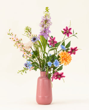 Het Zijden kunstbloemen boeket Bloom & Glow heeft roze, paarse, blauwe, oranje, witte en gele bloemen met groene bladeren in een roze vaas van keramiek tegen een lichte achtergrond.