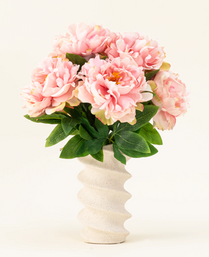 Het Zijden kunstbloemen boeket Peony Soft Pink heeft grote zachtroze pioenrozen en groene bladeren in een golvende, crèmekleurige vaas van keramiek, prachtig weergegeven tegen een effen lichte achtergrond.