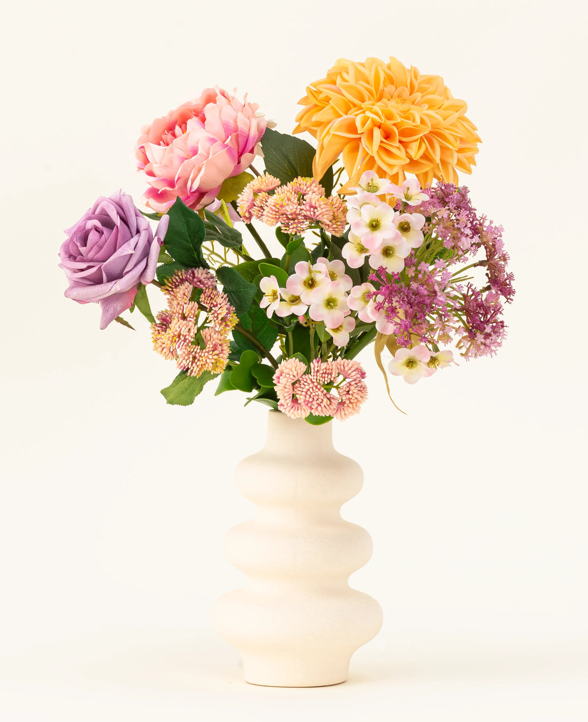 Un vase en céramique ondulée blanche présente le bouquet de fleurs artificielles en soie Peach Delight, comprenant un dahlia jaune, une pivoine rose, une rose violette et plusieurs petites fleurs blanches et roses sur un fond clair simple.