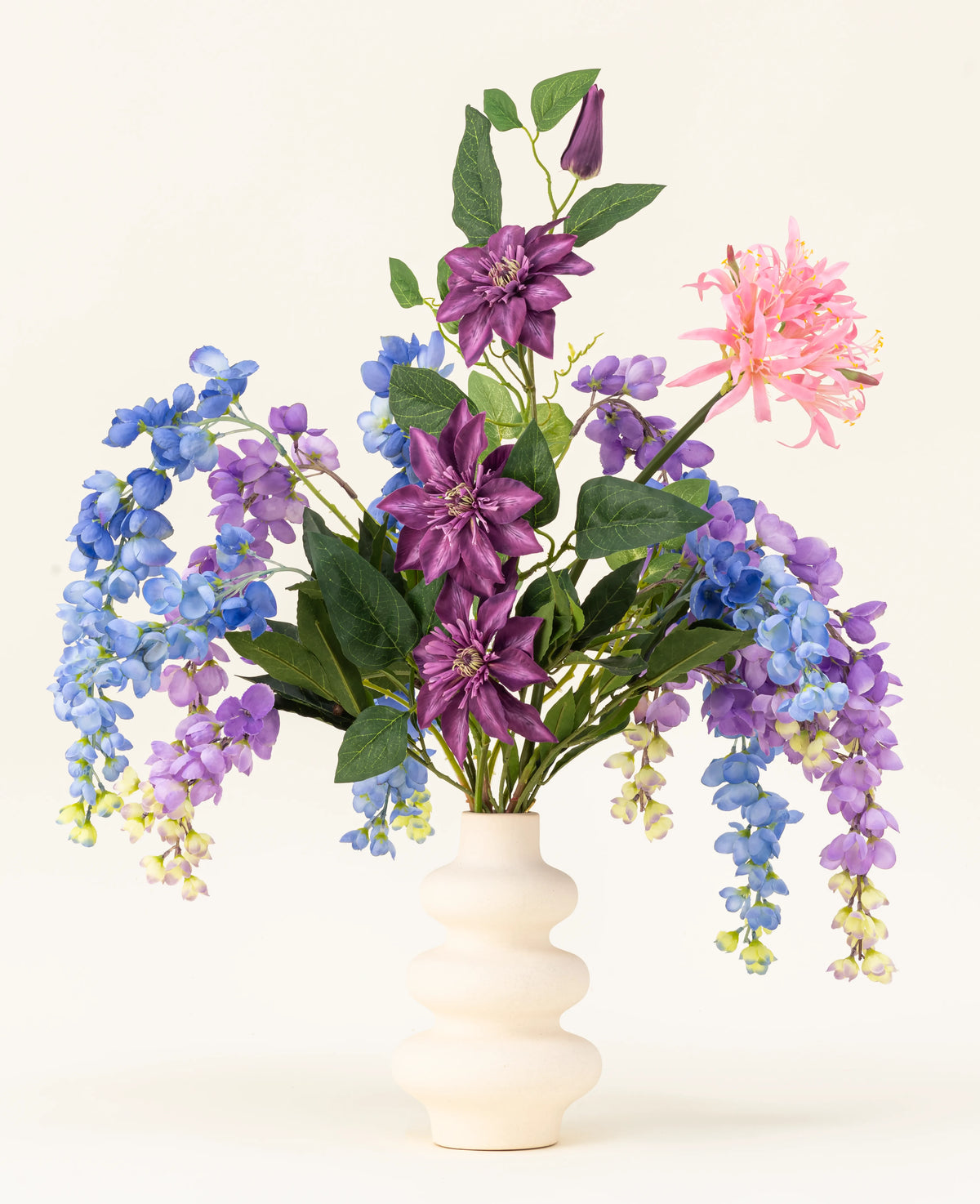 Un vase blanc ondulé présente le bouquet de fleurs artificielles en soie Indigo Dreams – un arrangement de fleurs artificielles violettes, bleues et roses avec des feuilles vertes – sur un fond clair uni.