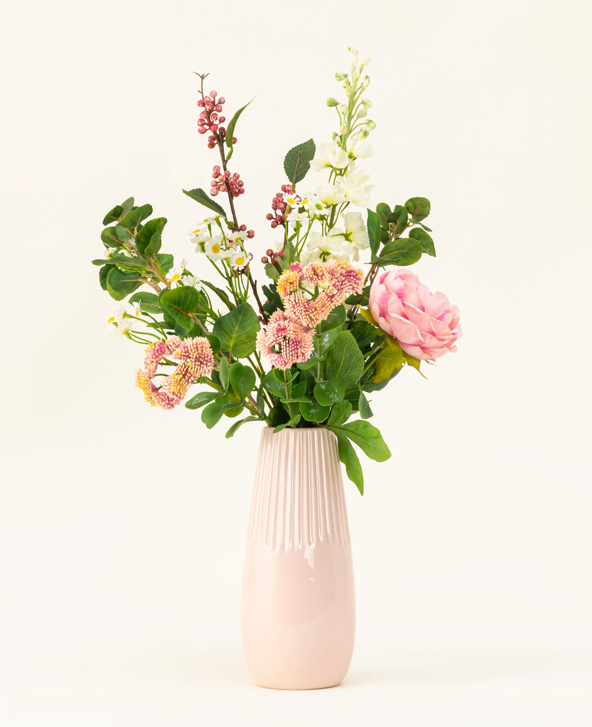 Un vase en céramique rose présente le bouquet de fleurs artificielles en soie Daisy Charm, avec diverses fleurs et verdure – roses roses, fleurs blanches et baies rouges – magnifiquement disposées sur un fond clair uni.