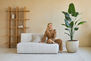 Een vrouw in een bruine outfit zit op een witte bank, glimlachend met een kopje in haar hand. Naast haar staat de Kunstplant Strelitzia 180 cm real touch, die weelderig groen brengt in de moderne, beige getinte woonkamer met minimale inrichting.