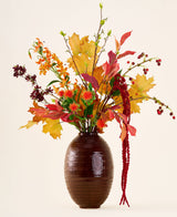Dans un vase marron se trouve le bouquet « Rustic Glow » de fleurs artificielles en soie, avec des feuilles jaunes, oranges et rouges, des baies rouges, des fleurs orange et de longues amarantes rouges pendantes sur un fond neutre.