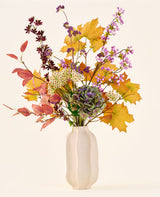 Le bouquet de fleurs artificielles en soie 'Wild Amethyst', avec des feuilles d'automne, des fleurs violettes et blanches et des hortensias verts, est présenté dans un vase en céramique blanche sur un fond uni et clair.