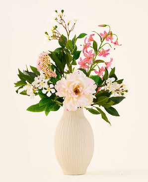 Dans un vase en céramique de couleur crème se trouve le bouquet « Light & Lovely » de fleurs artificielles en soie de Easyplants, composé d'un arrangement mixte de fleurs en soie blanches, roses et violet clair avec des feuilles vertes, sur un fond uni et clair.