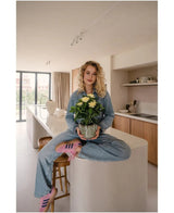 Une femme aux cheveux blonds bouclés, portant des vêtements en jean et des baskets roses, est assise les jambes croisées sur un îlot de cuisine dans une cuisine moderne et ensoleillée, tenant dans ses mains la plante artificielle Gardenia UV 39 cm.