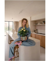 Une femme aux cheveux blonds bouclés, portant un jean et des chaussettes rayées, est assise souriante sur le comptoir de la cuisine tout en tenant la plante artificielle Gardenia UV 39 cm – une plante en pot à fleurs jaunes. En arrière-plan, vous pouvez voir une cuisine moderne avec des armoires et des étagères en bois.