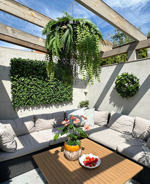 Une terrasse ensoleillée avec des sièges beiges avec des coussins et une table en bois, décorée d'un vase ananas à fleurs roses et d'un bol de fraises. Les plantes murales végétales offrent un intérieur luxuriant, complété par la fougère artificielle suspendue UV 60 cm avec anneau noir comme accent de suspension saisissant.