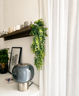Un espace de vie moderne avec une lampe en verre gris, des œuvres d'art encadrées, une plante en pot et une étagère en bois avec des tasses et la plante artificielle suspendue « Pannenkoek » (61 cm), dont les vrilles pendent à côté de rideaux blancs.