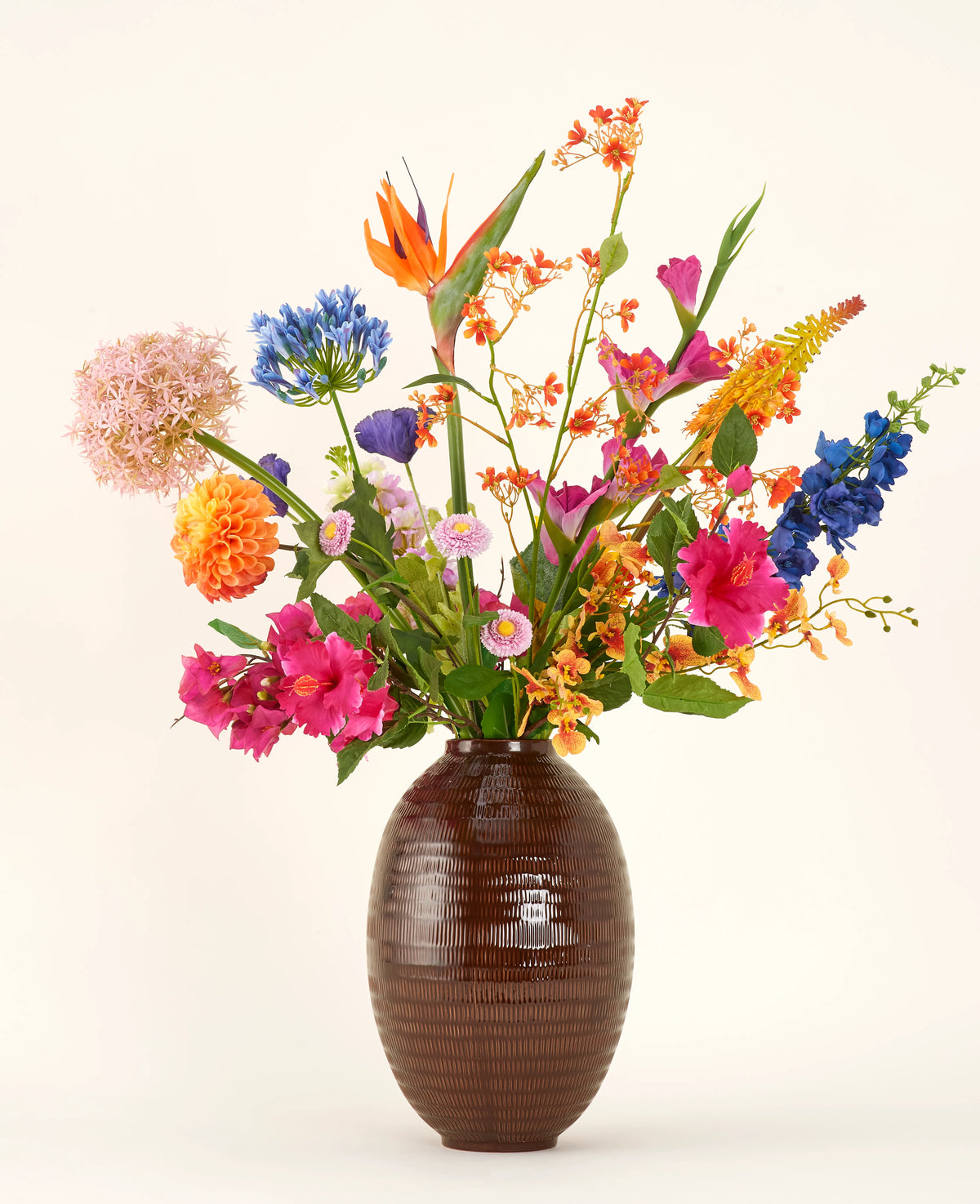 Dans un vase à structure marron se trouve le bouquet de fleurs artificielles en soie Tropical Firework - un arrangement de fleurs en soie orange, roses, violettes, jaunes et bleues avec des feuilles vertes sur un fond clair uni.