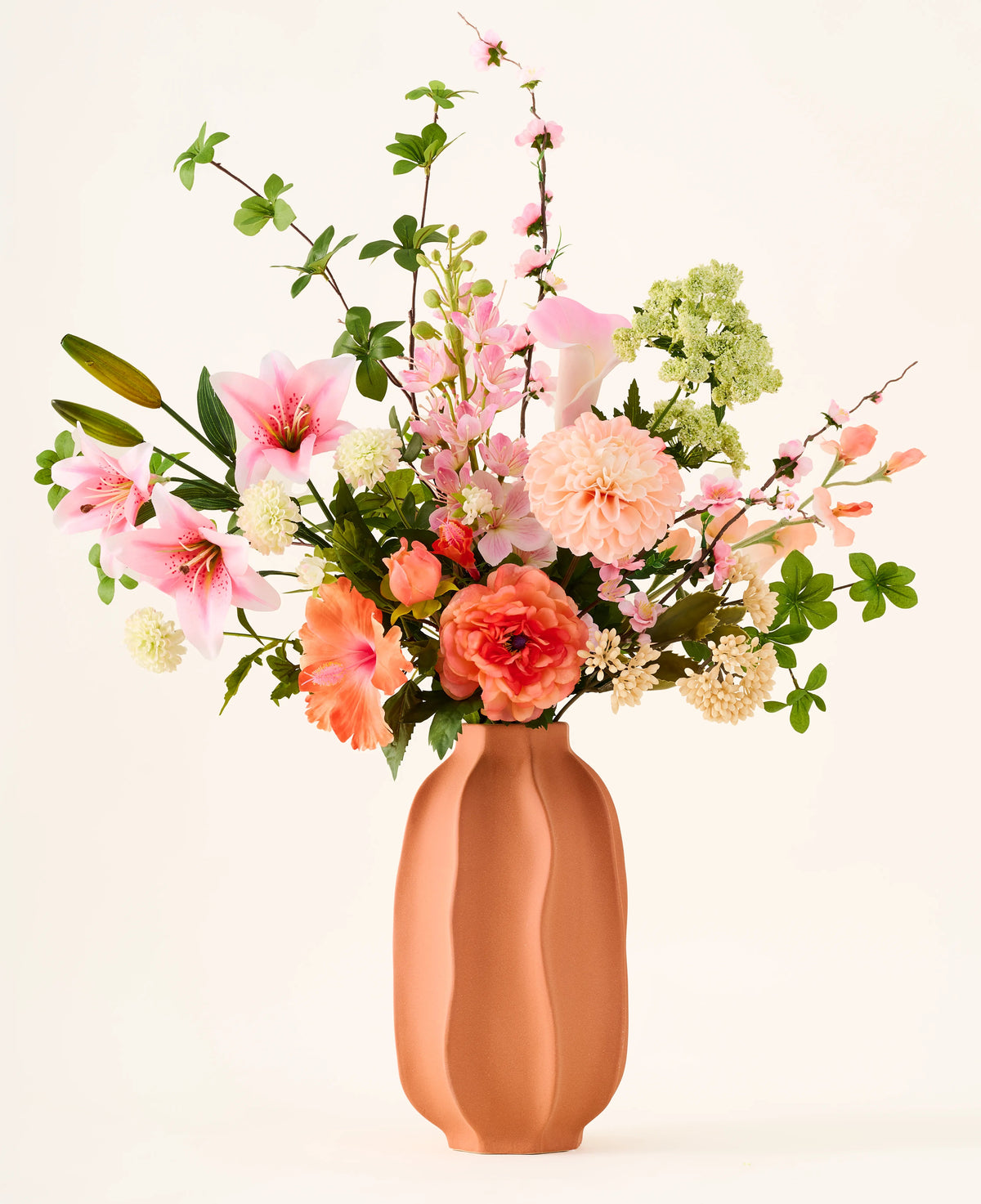 Dans un vase en terre cuite se trouve le bouquet coloré 'Yasmine's Choice' de fleurs artificielles en soie, composé de lys roses, de roses pêche et de feuilles vertes, composé de fleurs en soie de haute qualité d'Easyplants, le tout magnifiquement disposé sur un fond clair.
