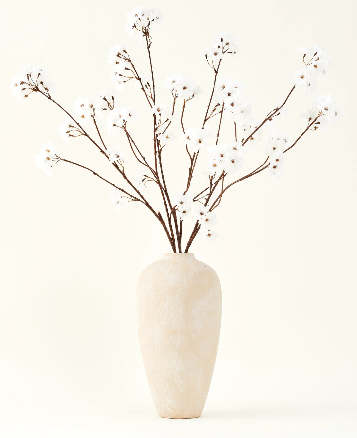 Le bouquet de fleurs artificielles en soie Fleur de cerisier crème 114 cm se dresse haut dans un vase en céramique beige et apporte une décoration sereine à n'importe quelle pièce avec ses délicates branches de couleur crème sur un fond clair.