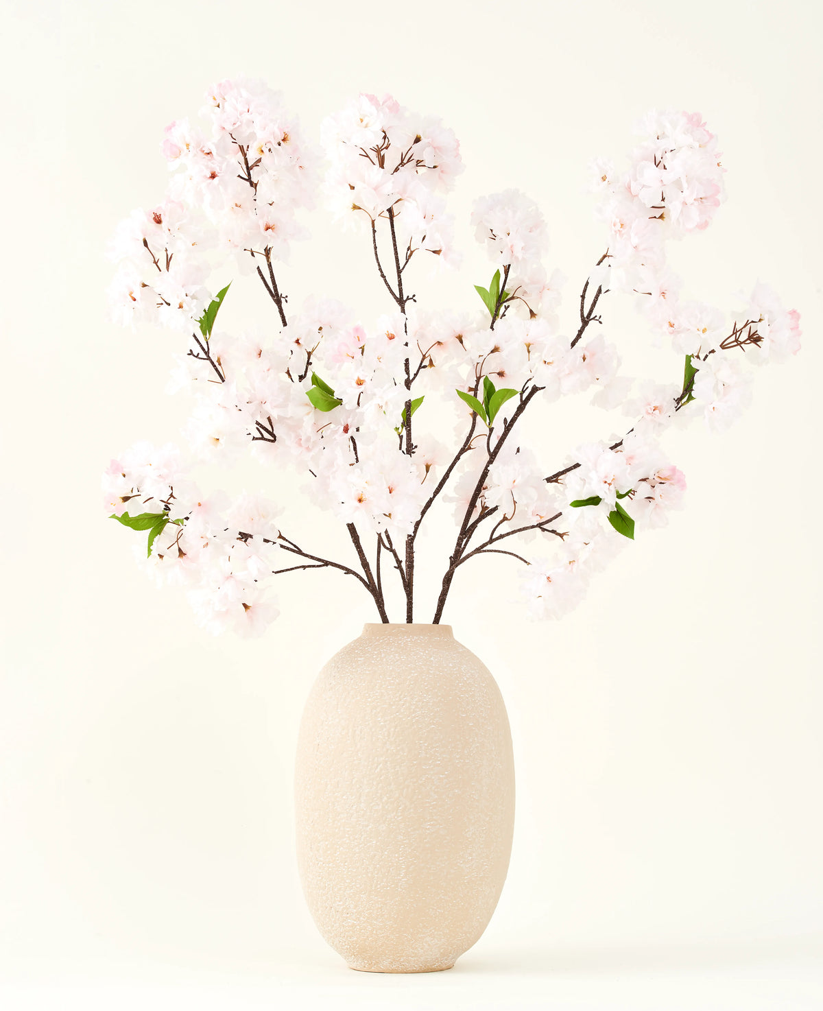 Le bouquet de fleurs artificielles en soie Cherry Blossom Blush 119 cm présente de délicates fleurs de cerisier rose clair avec des feuilles vertes dans un vase beige, idéal comme décoration de fleurs artificielles sur un fond clair.