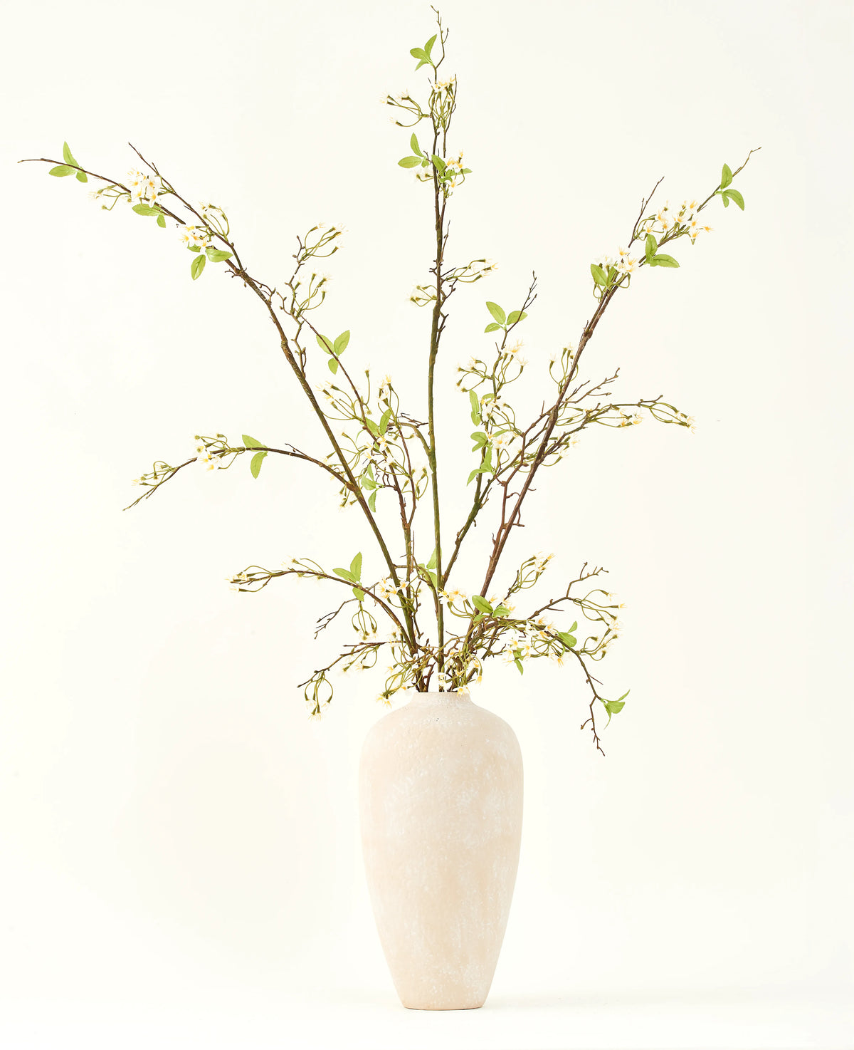 Un grand vase en céramique de couleur crème contient le bouquet de fleurs artificielles en soie Verveine crème avec de longues branches, de petites feuilles vertes et de délicates fleurs blanches, joliment disposées sur un fond clair et uni.