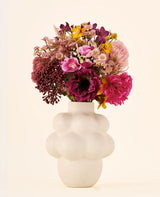 Un bouquet de fleurs artificielles en soie, A Touch of Ashley, dans les tons rose, violet et jaune, présenté dans un vase blanc cassé en forme de bulle et à surface structurée.