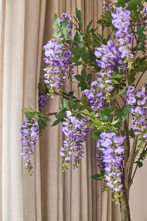Voor de beige gordijnen hangt een kunstplant Blauweregen van 150 cm met groene bladeren.