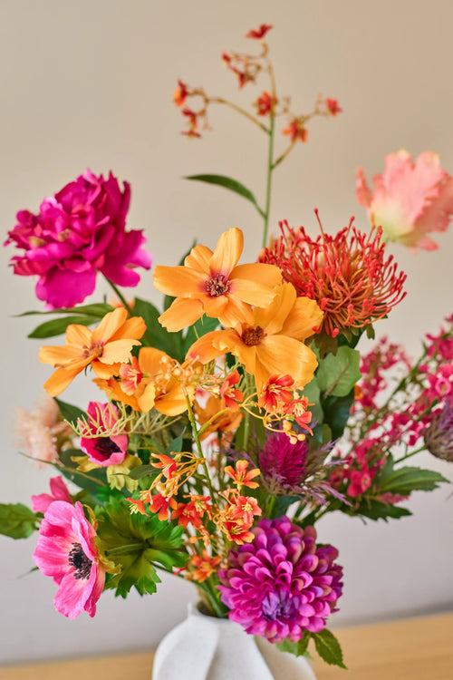 Un bouquet vibrant de fleurs artificielles de Zijden, « Magnus di Lambo », composé d'un mélange de fleurs artificielles orange, roses et magenta, est disposé dans un vase blanc sur un fond neutre.