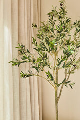 De Kunst Olijfboom van 180 cm, met weelderig groen blad en realistische zwarte olijven, staat voor beige gordijnen en een neutraalgekleurde muur. Zonlicht verlicht het tafereel zachtjes en geeft de ruimte een vleugje mediterrane stijl.