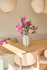 Op een lichte houten tafel in een lichte, moderne eethoek met neutrale tinten en zacht zonlicht dat naar binnen stroomt, staat een witte vaas met het boeket ‘Purple Dawn’ van Zijden kunstbloemen.