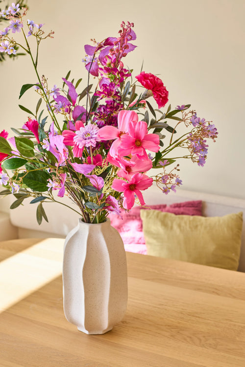 Dans un vase en céramique blanche se trouve le bouquet de fleurs artificielles en soie « Purple Dawn », un arrangement vivant de fleurs roses, violettes et magenta ; le vase repose sur une table en bois clair avec des coussins colorés en arrière-plan.