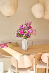 Op een lichte houten eettafel staat een witte vaas met het boeket ‘Purple Dawn’ van Zijden kunstbloemen – een prachtig boeket van zijden bloemen in roze en paarse tinten. Beige stoelen, een ingebouwde bank met kussens en grote papieren lantaarns maken de gezellige, zonovergoten kamer compleet.