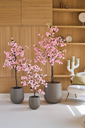 Drie potplanten van het type Kunstplant Bougainvillea van 120 cm, roze van kleur en met roze bloemen, staan op een witte vloer in een moderne kamer met houten lambrisering, planken en een witte stoel.