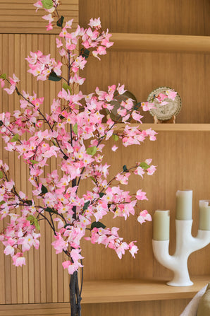 Een kunstplant bougainvillea van 150 cm, roze van kleur en met roze bloesems, staat voor houten planken waarop een decoratief bord en een moderne witte kandelaar met drie groene kaarsen staan uitgestald.
